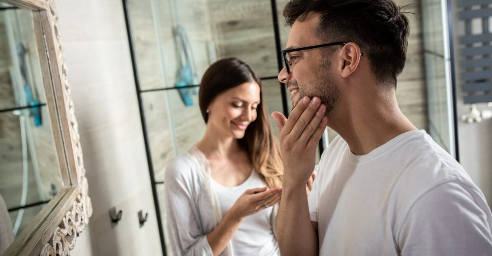 Man applying facial skincare cream in bathroom to help prevent ingrown hair cysts, with partner smiling in background